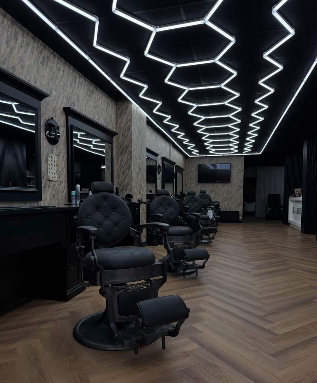 Zwarte barberstoelen op een visgraatvloer onder een plafond van witte lichtlijnen